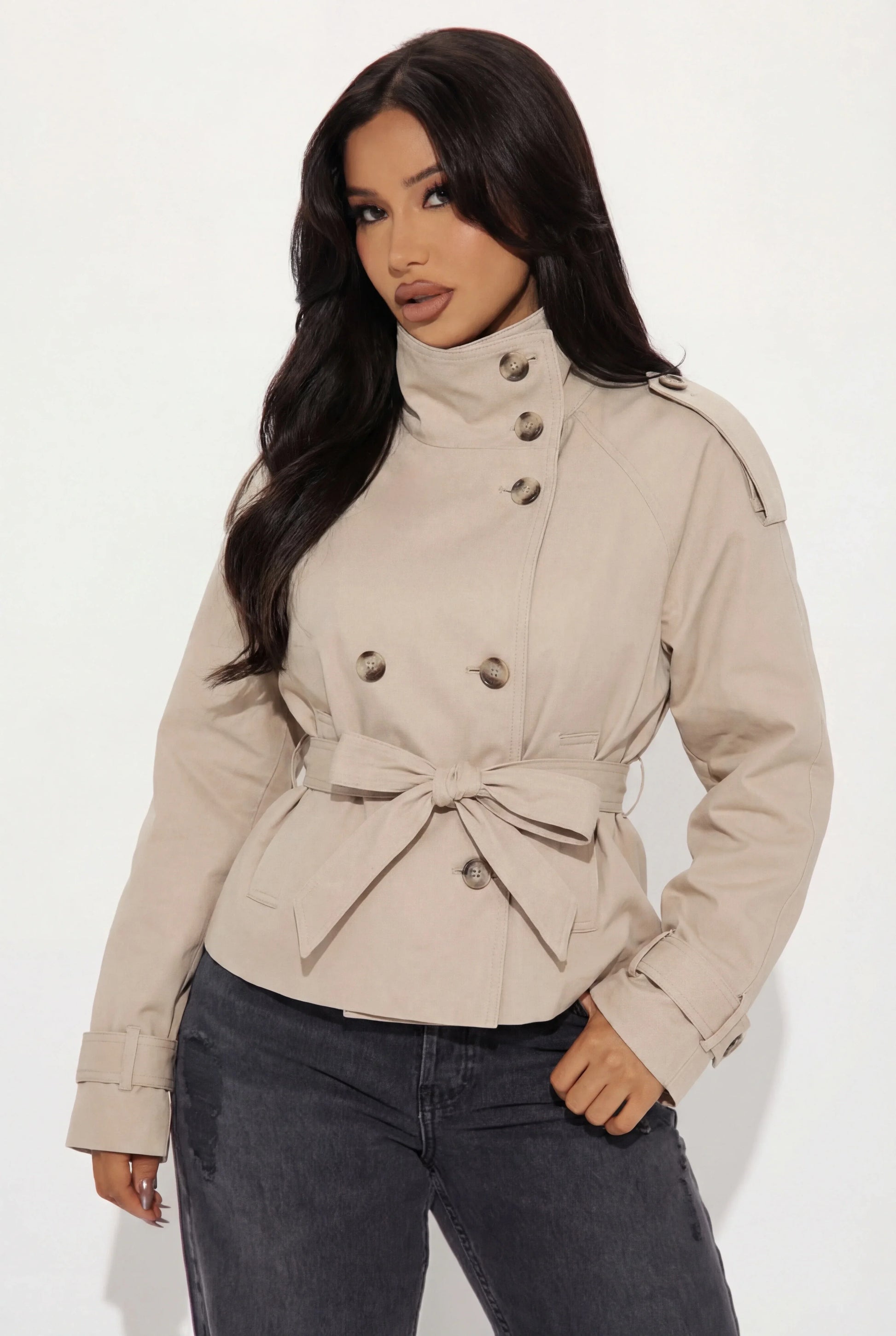 Beige - A woman wearing a beige cropped twill trench jacket with a high funnel neck, double-breasted button closure, and a cinched self-tie waist belt, paired with dark grey jeans.