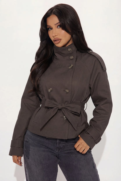 Brown - A woman wearing a beige cropped twill trench jacket with a high funnel neck, double-breasted button closure, and a cinched self-tie waist belt, paired with dark grey jeans.