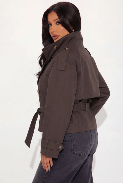 Brown - A woman wearing a beige cropped twill trench jacket with a high funnel neck, double-breasted button closure, and a cinched self-tie waist belt, paired with dark grey jeans.