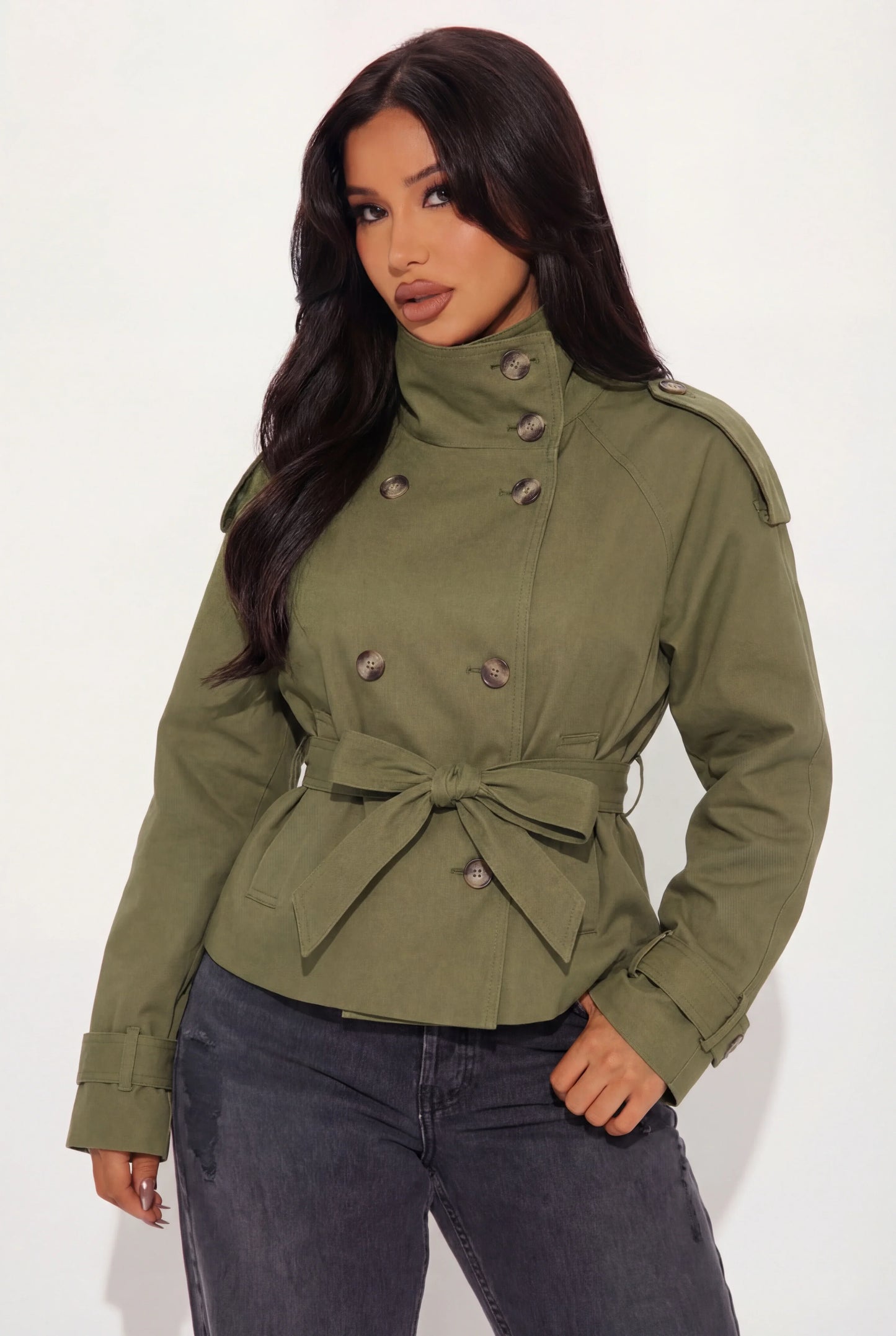 Khaki - A woman wearing a beige cropped twill trench jacket with a high funnel neck, double-breasted button closure, and a cinched self-tie waist belt, paired with dark grey jeans.