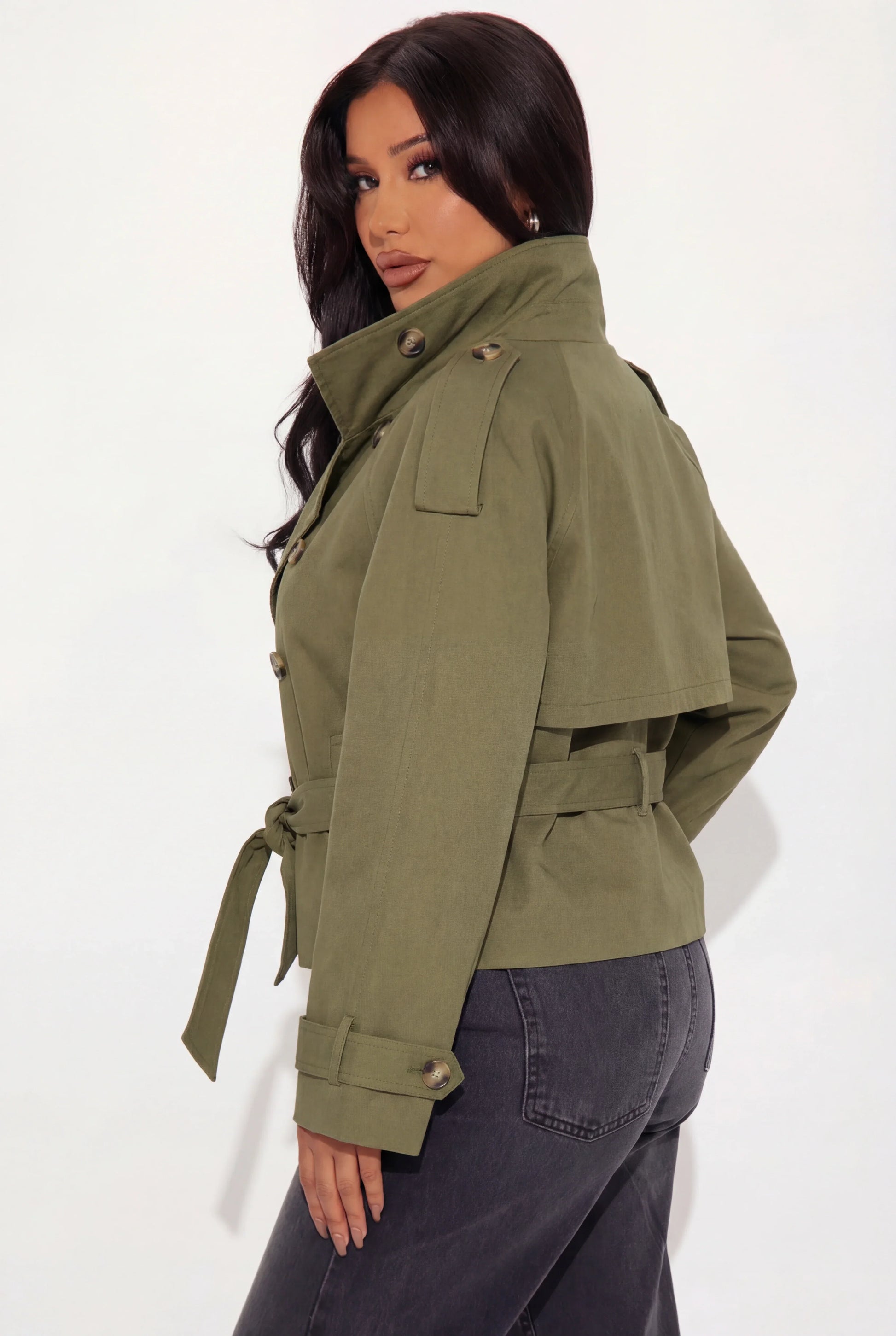 Khaki - A woman wearing a beige cropped twill trench jacket with a high funnel neck, double-breasted button closure, and a cinched self-tie waist belt, paired with dark grey jeans.