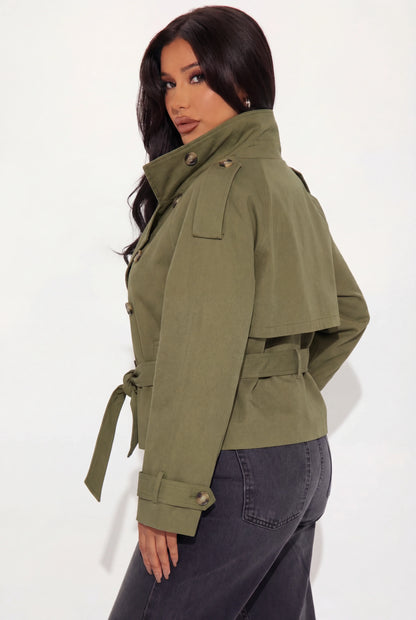Khaki - A woman wearing a beige cropped twill trench jacket with a high funnel neck, double-breasted button closure, and a cinched self-tie waist belt, paired with dark grey jeans.