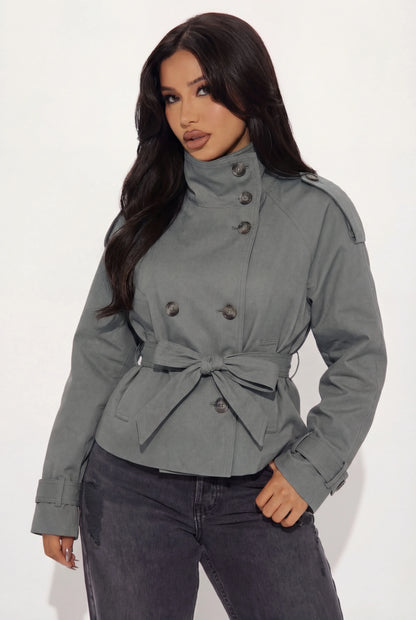 Grey - A woman wearing a beige cropped twill trench jacket with a high funnel neck, double-breasted button closure, and a cinched self-tie waist belt, paired with dark grey jeans.