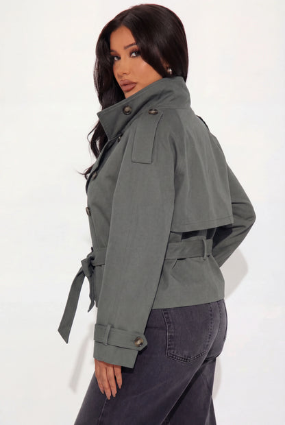 Grey - A woman wearing a beige cropped twill trench jacket with a high funnel neck, double-breasted button closure, and a cinched self-tie waist belt, paired with dark grey jeans.