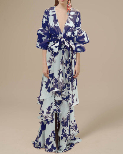 Blue - A woman wearing a pale blue and mustard yellow botanical print maxi dress featuring a deep V-neckline, voluminous balloon sleeves, a belted waist, and a cascading tiered ruffle skirt.