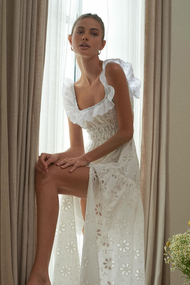 White - A woman wearing a white broderie anglaise maxi dress with a ruffled neckline, smocked waist, and a high thigh slit.