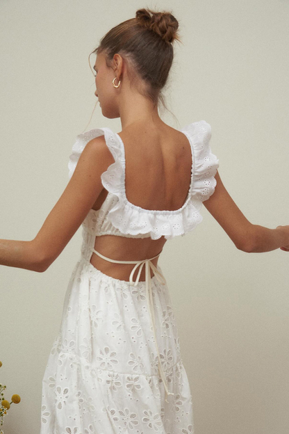 White - A woman wearing a white broderie anglaise maxi dress with a ruffled neckline, smocked waist, and a high thigh slit.