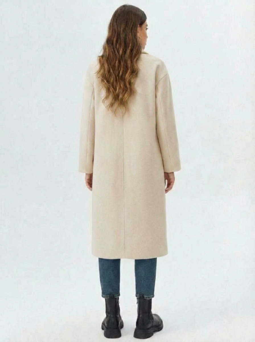 Beige - A woman wearing a camel-coloured, double-breasted longline coat with notch lapels and folded cuffs, styled over a striped sweater, blue jeans, and black boots against a white background.