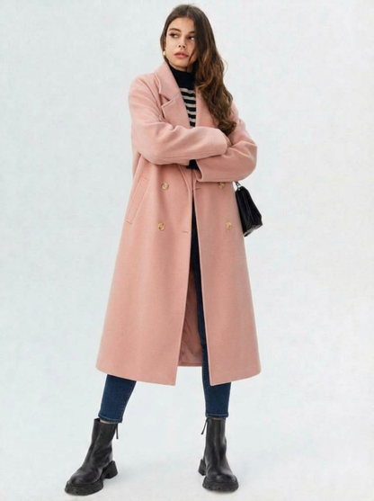 Pink - A woman wearing a camel-coloured, double-breasted longline coat with notch lapels and folded cuffs, styled over a striped sweater, blue jeans, and black boots against a white background.
