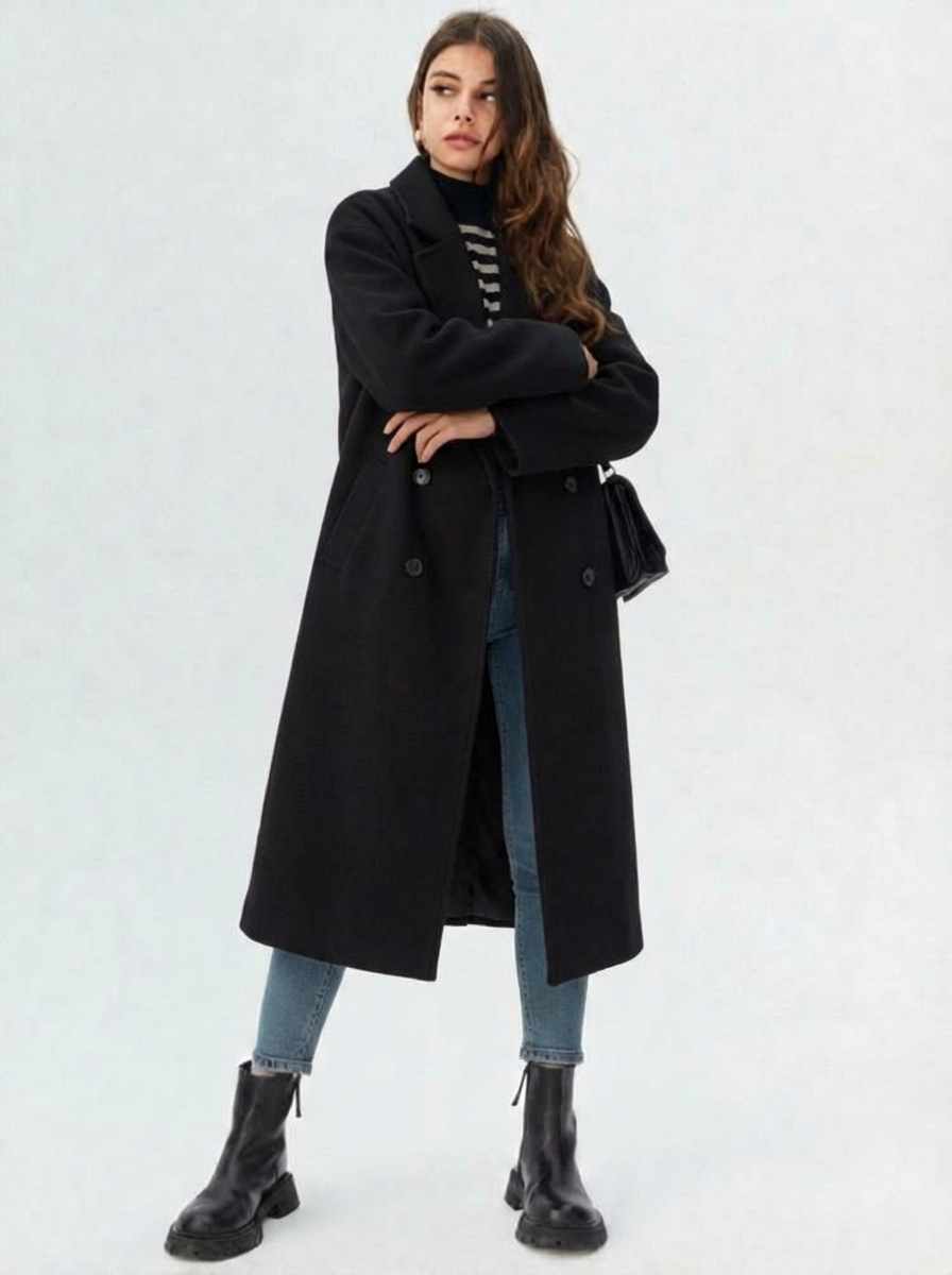 Black - A woman wearing a camel-coloured, double-breasted longline coat with notch lapels and folded cuffs, styled over a striped sweater, blue jeans, and black boots against a white background.