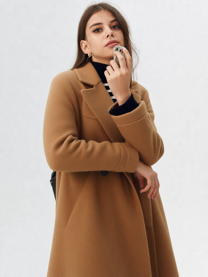 A woman wearing a camel-coloured, double-breasted longline coat with notch lapels and folded cuffs, styled over a striped sweater, blue jeans, and black boots against a white background.