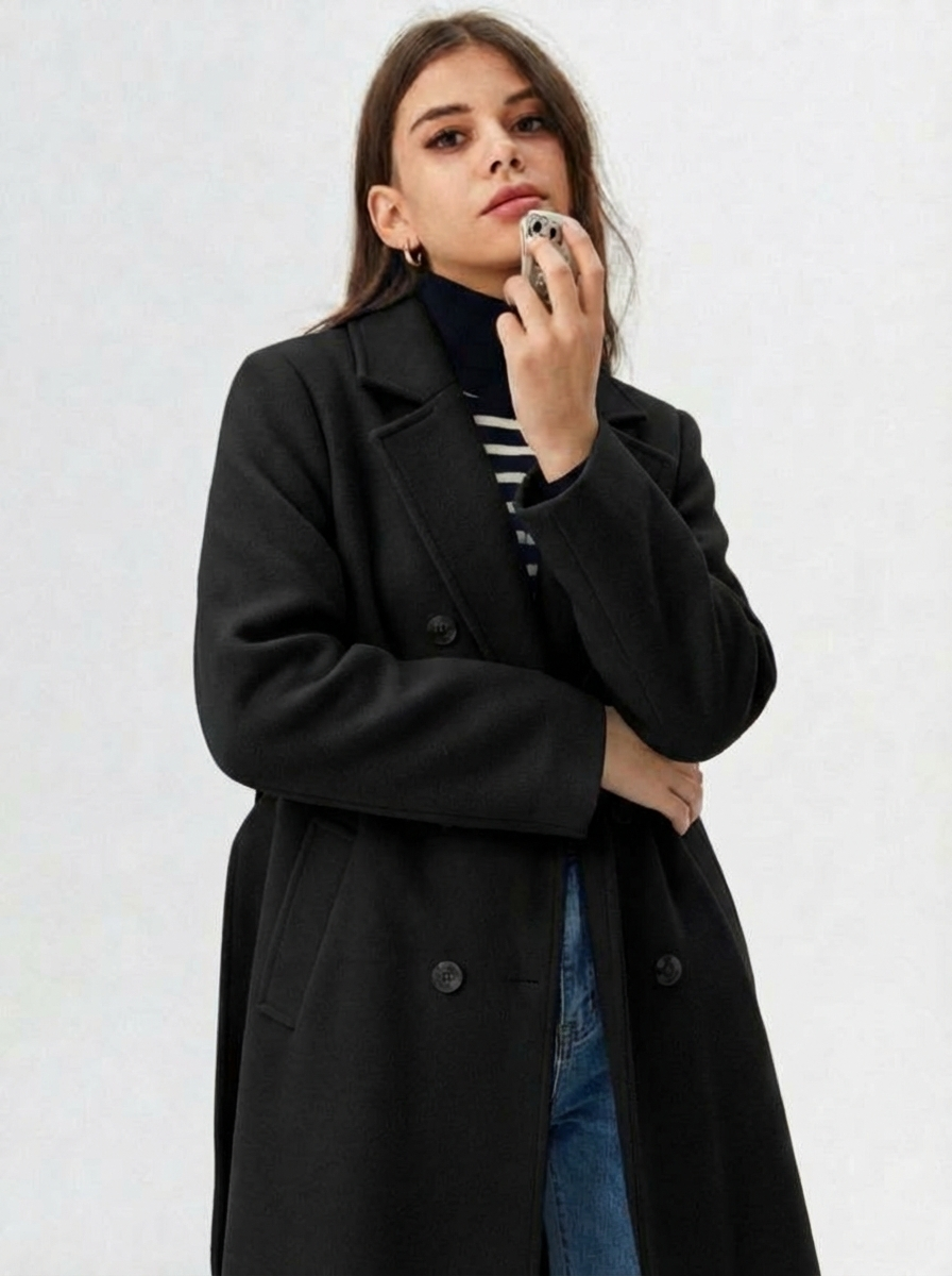 Black - A woman wearing a camel-coloured, double-breasted longline coat with notch lapels and folded cuffs, styled over a striped sweater, blue jeans, and black boots against a white background.