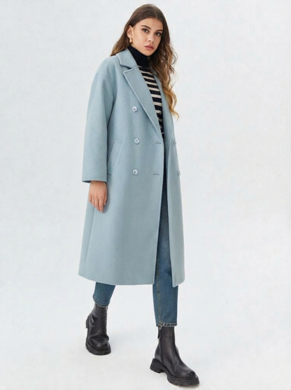 Blue - A woman wearing a camel-coloured, double-breasted longline coat with notch lapels and folded cuffs, styled over a striped sweater, blue jeans, and black boots against a white background.