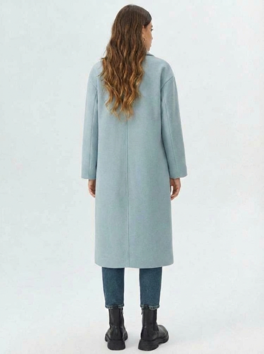 Blue - A woman wearing a camel-coloured, double-breasted longline coat with notch lapels and folded cuffs, styled over a striped sweater, blue jeans, and black boots against a white background.