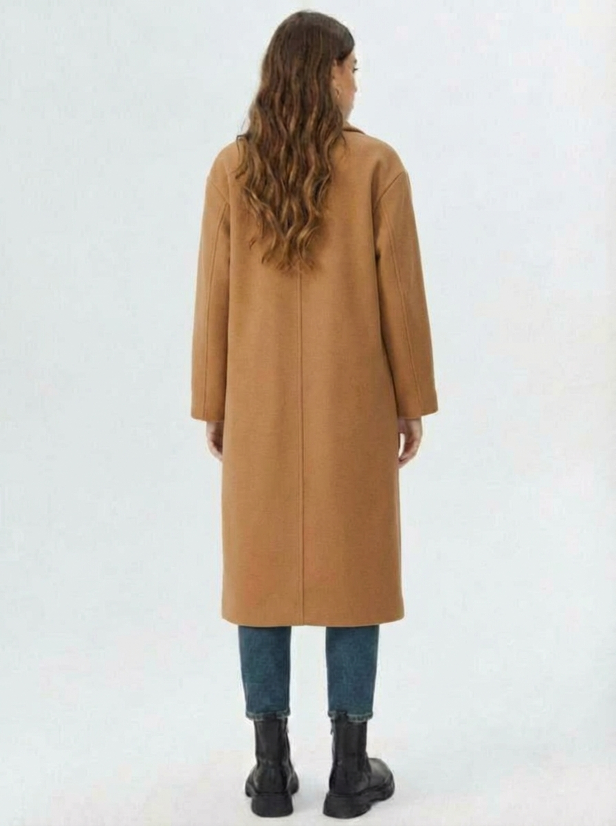 Brown - A woman wearing a camel-coloured, double-breasted longline coat with notch lapels and folded cuffs, styled over a striped sweater, blue jeans, and black boots against a white background.