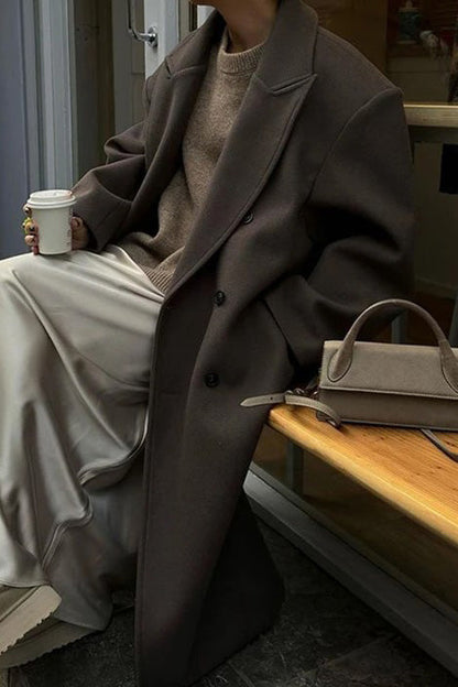 Brown - A person wearing a longline, double-breasted dark chocolate brown overcoat with notch lapels, styled over a beige knit sweater and cream silk skirt.