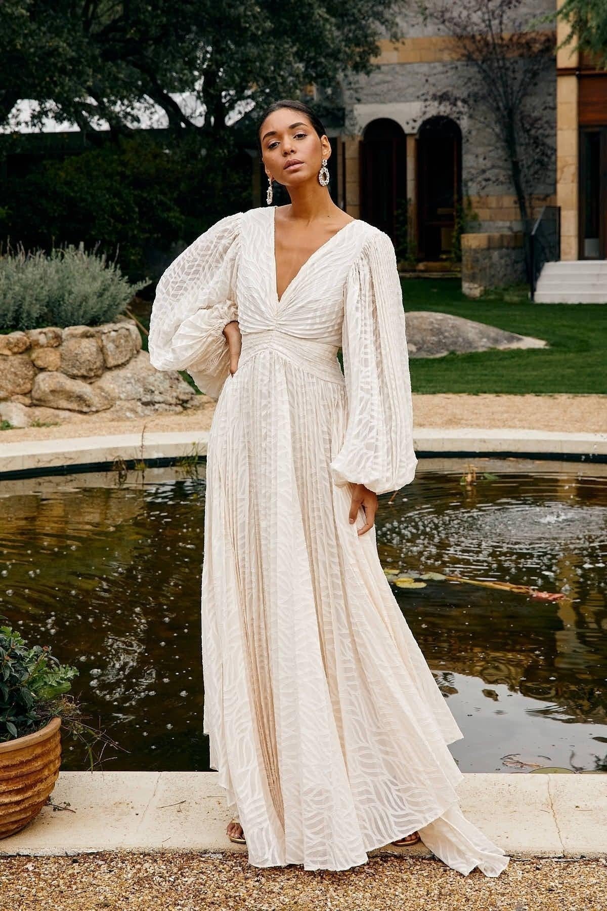 WHite - A woman stands outdoors near a pond wearing a long, flowing white maxi dress with a deep V-neck, ruched bodice, voluminous long sleeves, and an intricate burnout texture pattern.