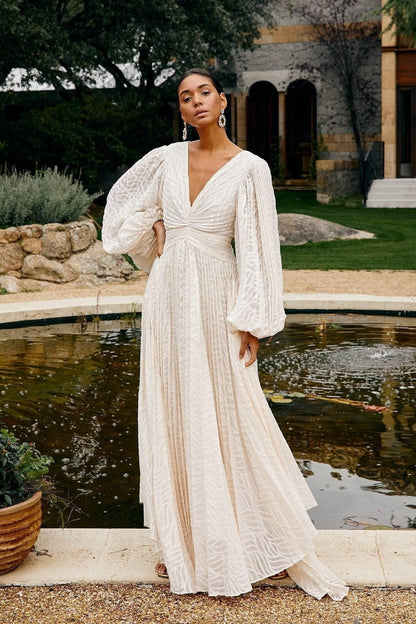 WHite - A woman stands outdoors near a pond wearing a long, flowing white maxi dress with a deep V-neck, ruched bodice, voluminous long sleeves, and an intricate burnout texture pattern.