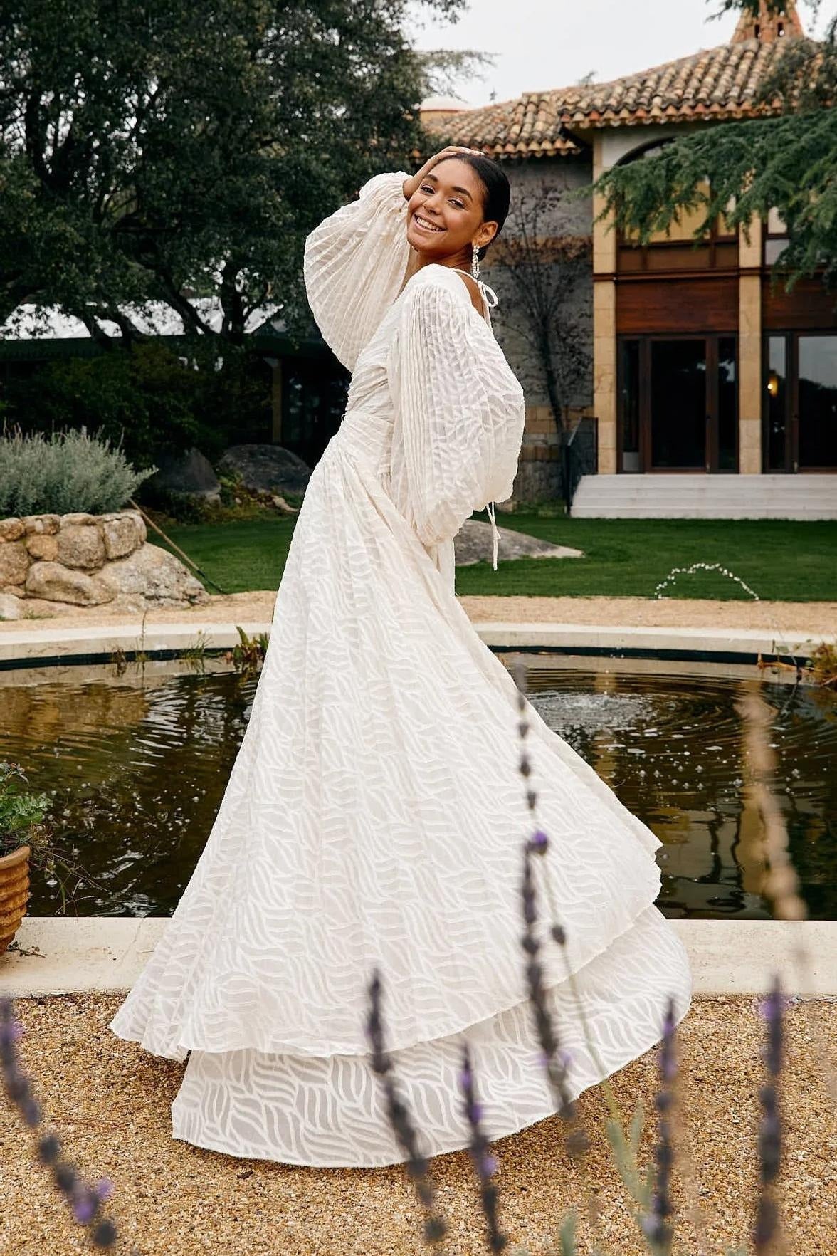 WHite - A woman stands outdoors near a pond wearing a long, flowing white maxi dress with a deep V-neck, ruched bodice, voluminous long sleeves, and an intricate burnout texture pattern.
