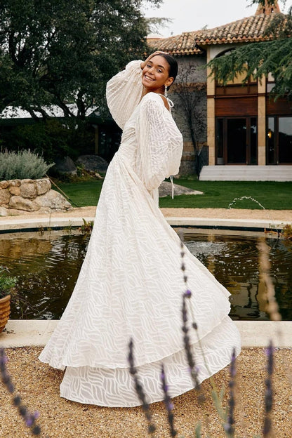 WHite - A woman stands outdoors near a pond wearing a long, flowing white maxi dress with a deep V-neck, ruched bodice, voluminous long sleeves, and an intricate burnout texture pattern.