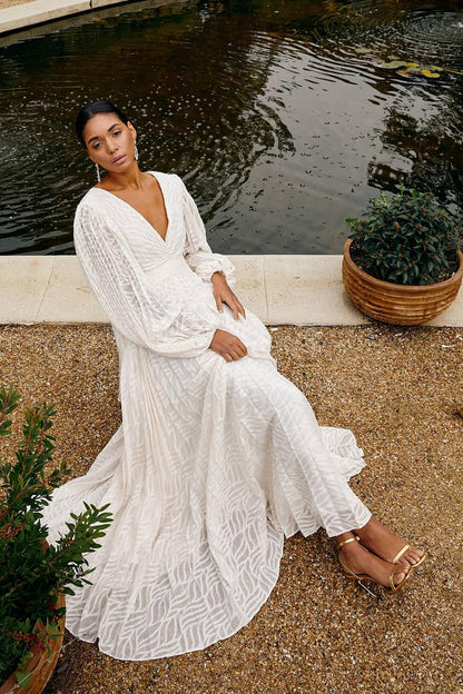 WHite - A woman stands outdoors near a pond wearing a long, flowing white maxi dress with a deep V-neck, ruched bodice, voluminous long sleeves, and an intricate burnout texture pattern.