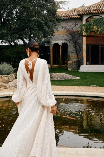 WHite - A woman stands outdoors near a pond wearing a long, flowing white maxi dress with a deep V-neck, ruched bodice, voluminous long sleeves, and an intricate burnout texture pattern.