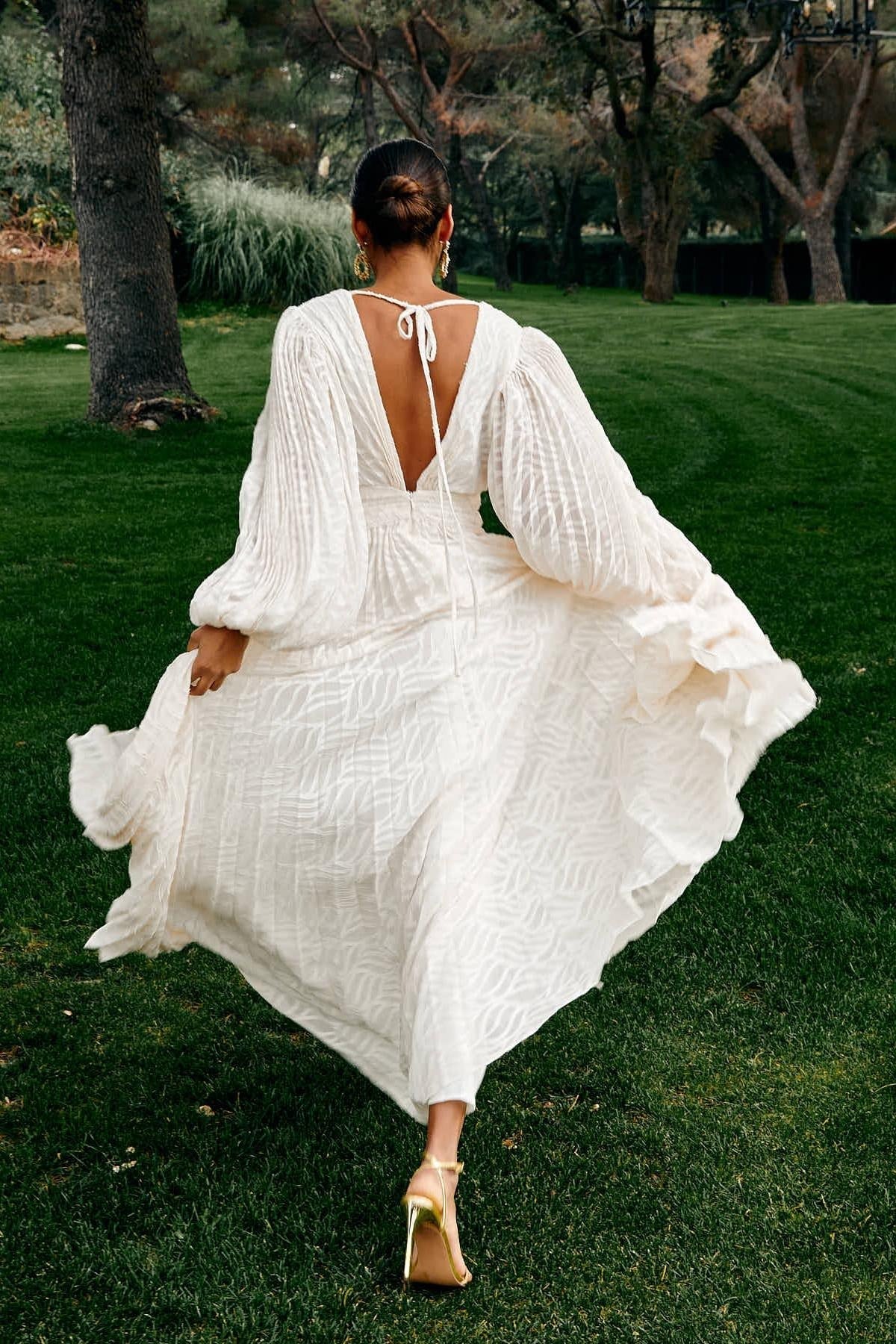 WHite - A woman stands outdoors near a pond wearing a long, flowing white maxi dress with a deep V-neck, ruched bodice, voluminous long sleeves, and an intricate burnout texture pattern.