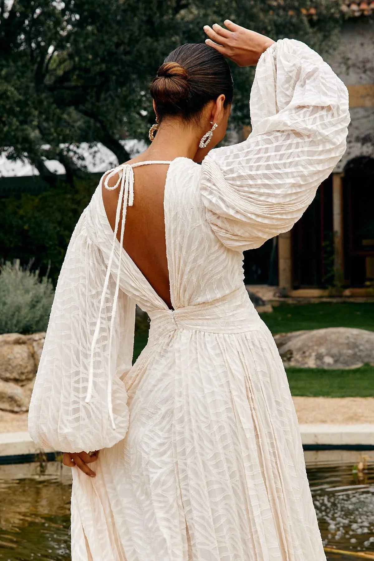 WHite - A woman stands outdoors near a pond wearing a long, flowing white maxi dress with a deep V-neck, ruched bodice, voluminous long sleeves, and an intricate burnout texture pattern.