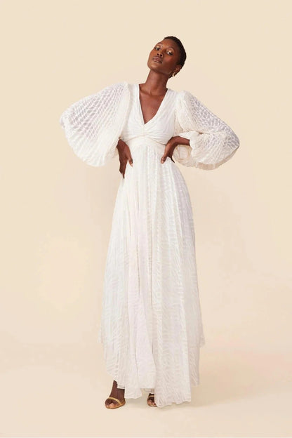 WHite - A woman stands outdoors near a pond wearing a long, flowing white maxi dress with a deep V-neck, ruched bodice, voluminous long sleeves, and an intricate burnout texture pattern.