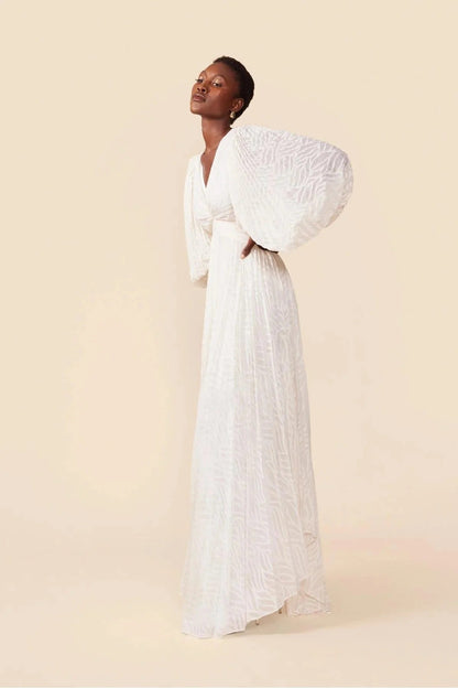 WHite - A woman stands outdoors near a pond wearing a long, flowing white maxi dress with a deep V-neck, ruched bodice, voluminous long sleeves, and an intricate burnout texture pattern.