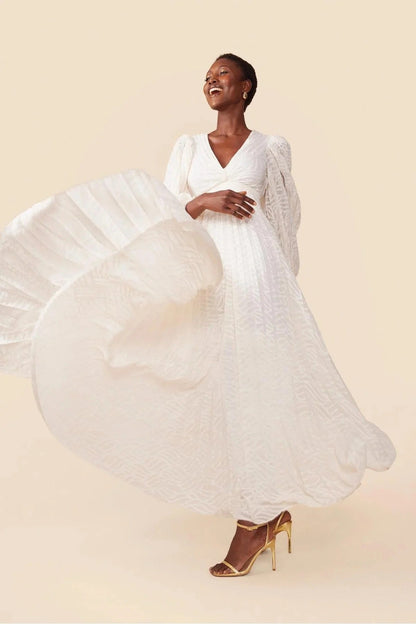 WHite - A woman stands outdoors near a pond wearing a long, flowing white maxi dress with a deep V-neck, ruched bodice, voluminous long sleeves, and an intricate burnout texture pattern.