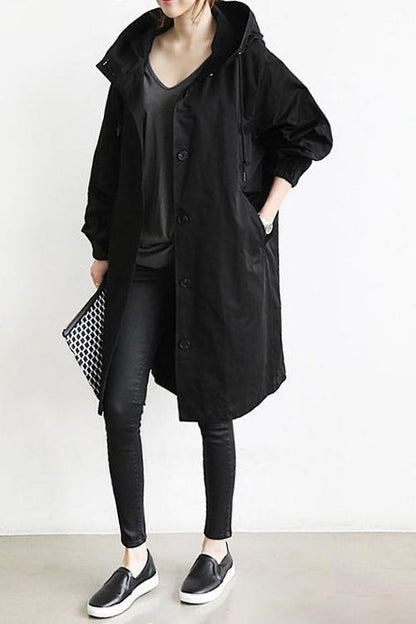 Pink - A woman wearing a teal mid-length hooded coat with a contrast black button-down front, angled flap pockets, and elasticated cuffs, set against a bright minimalist interior.
