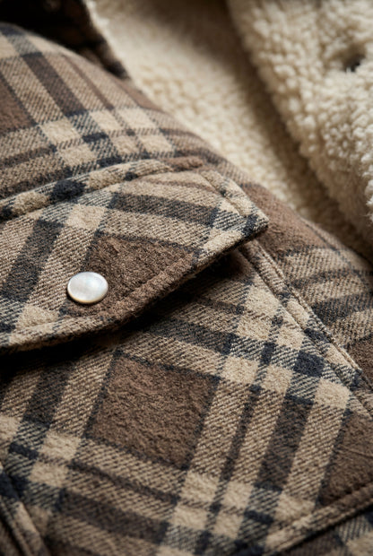 Navy Blue - A woman wearing a brown plaid flannel shirt jacket with a cream shearling lining, paired with dark wash jeans and brown leather boots.