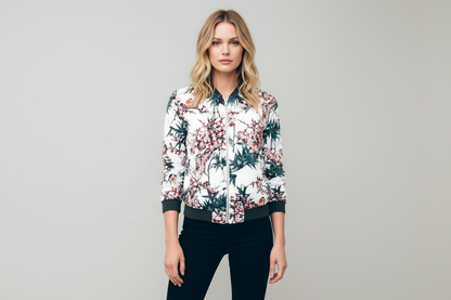 White - A woman wearing a white bomber jacket featuring a detailed pink cherry blossom and green leaf print, styled with black pants against a neutral grey background.