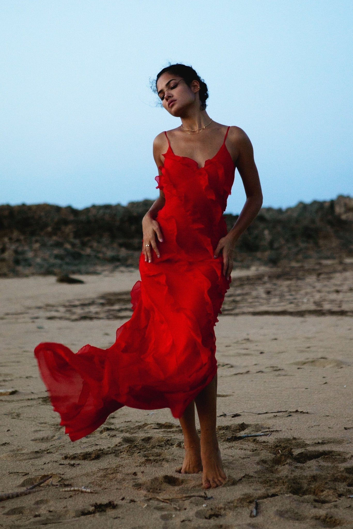 Red - A woman wearing a vibrant red chiffon maxi dress with cascading diagonal ruffles, a V-neckline, and thin spaghetti straps, posing against a rustic dark wooden door.