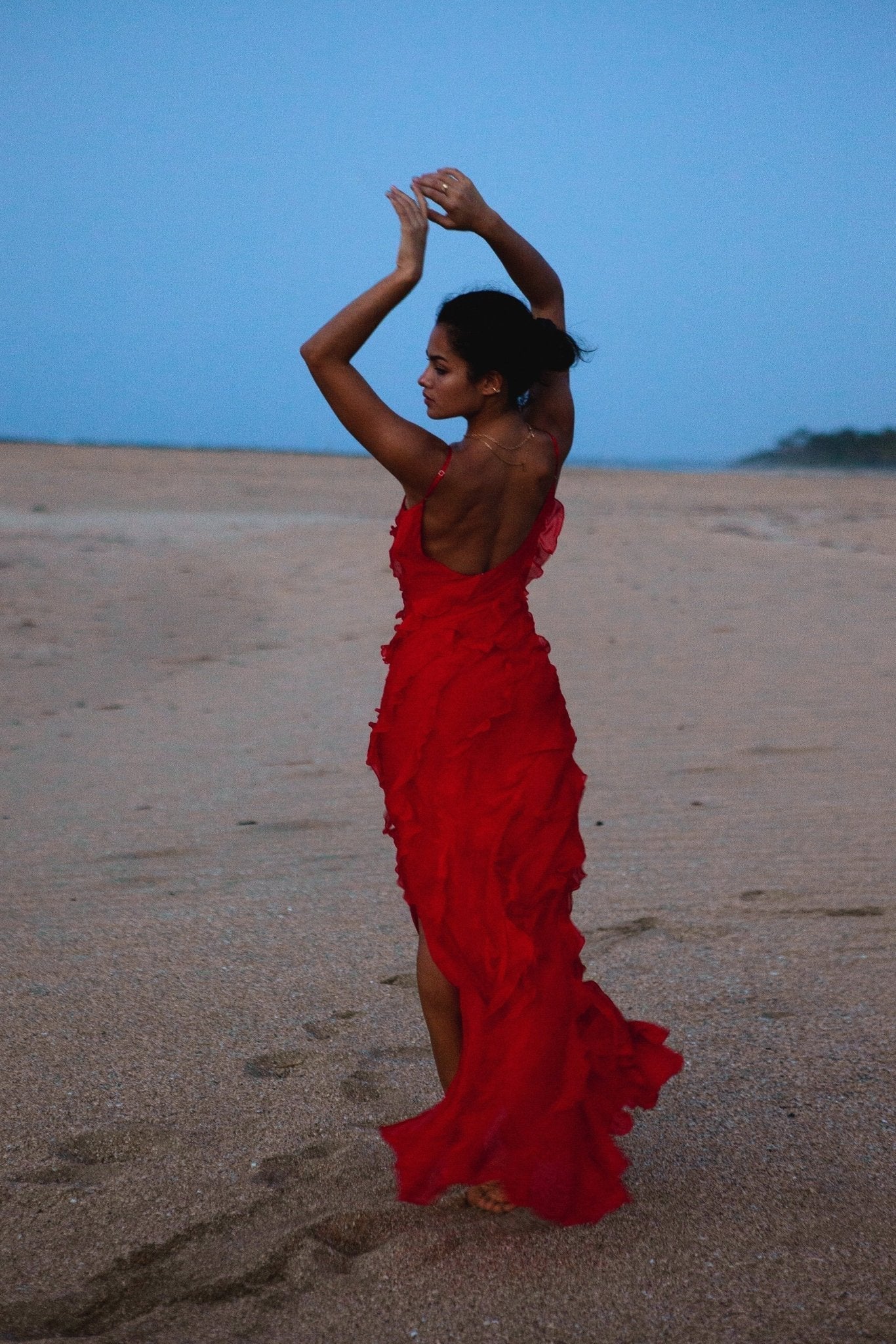 Red - A woman wearing a vibrant red chiffon maxi dress with cascading diagonal ruffles, a V-neckline, and thin spaghetti straps, posing against a rustic dark wooden door.