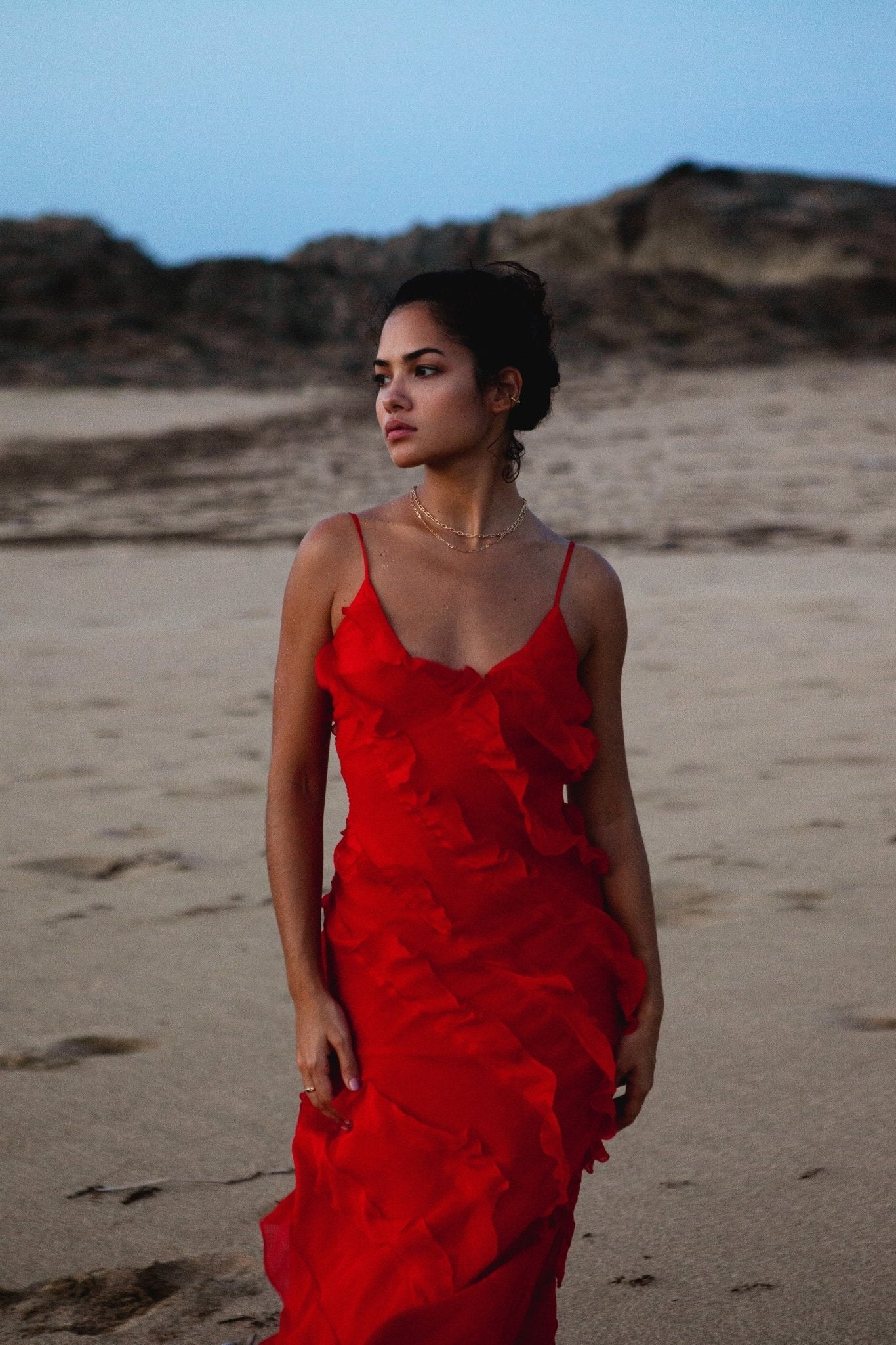 Red - A woman wearing a vibrant red chiffon maxi dress with cascading diagonal ruffles, a V-neckline, and thin spaghetti straps, posing against a rustic dark wooden door.