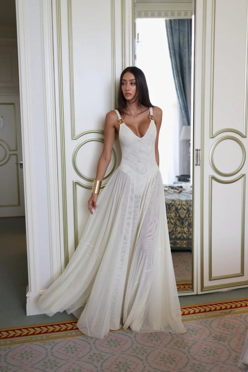 White - A woman wearing an elegant off-white maxi dress featuring a structured plunging bodice with gold hardware shoulder straps and a sheer, flowing pleated chiffon skirt.