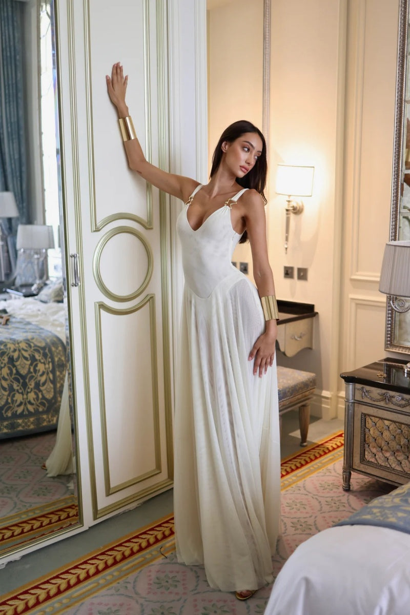 White - A woman wearing an elegant off-white maxi dress featuring a structured plunging bodice with gold hardware shoulder straps and a sheer, flowing pleated chiffon skirt.