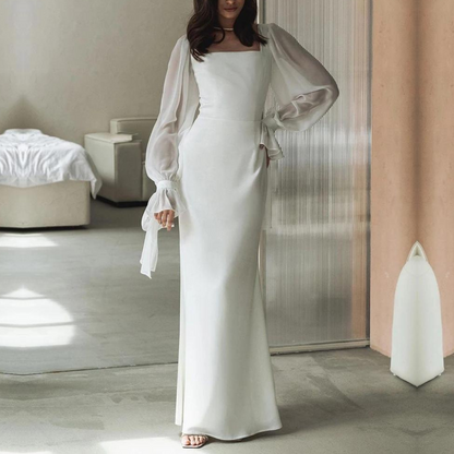 A woman wearing an elegant floor-length white crepe maxi dress featuring a square neckline and sheer, voluminous chiffon balloon sleeves with tied cuffs.