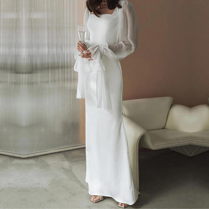 A woman wearing an elegant floor-length white crepe maxi dress featuring a square neckline and sheer, voluminous chiffon balloon sleeves with tied cuffs.