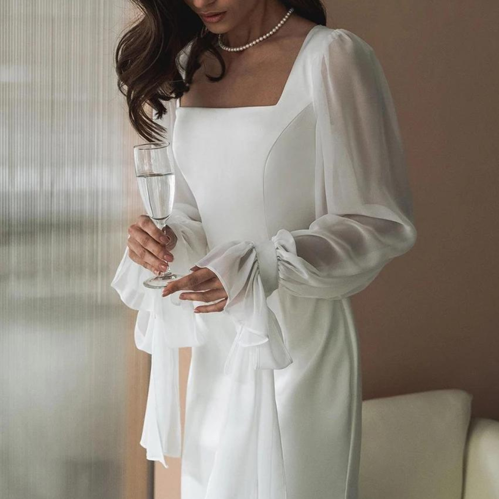 A woman wearing an elegant floor-length white crepe maxi dress featuring a square neckline and sheer, voluminous chiffon balloon sleeves with tied cuffs.