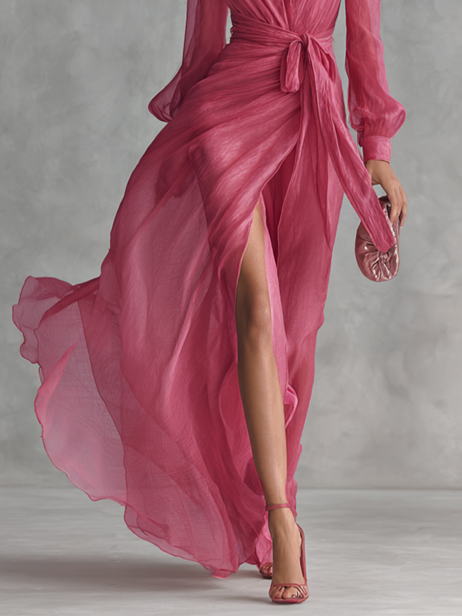 Pink - A woman wearing a rose pink long-sleeve chiffon maxi dress with a plunging V-neckline, a tied waist belt, and a high leg split, walking forward as the lightweight fabric drapes and flows.