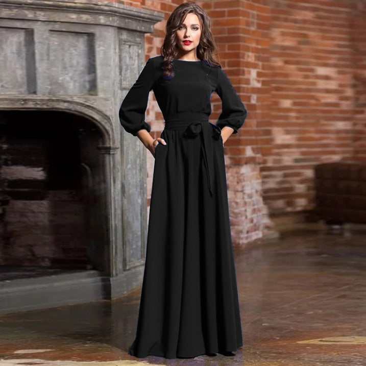 Black - A woman posing in a floor-length white crepe maxi dress featuring a boat neckline, three-quarter balloon sleeves, and a self-tie fabric sash at the waist, set against a modern interior backdrop.