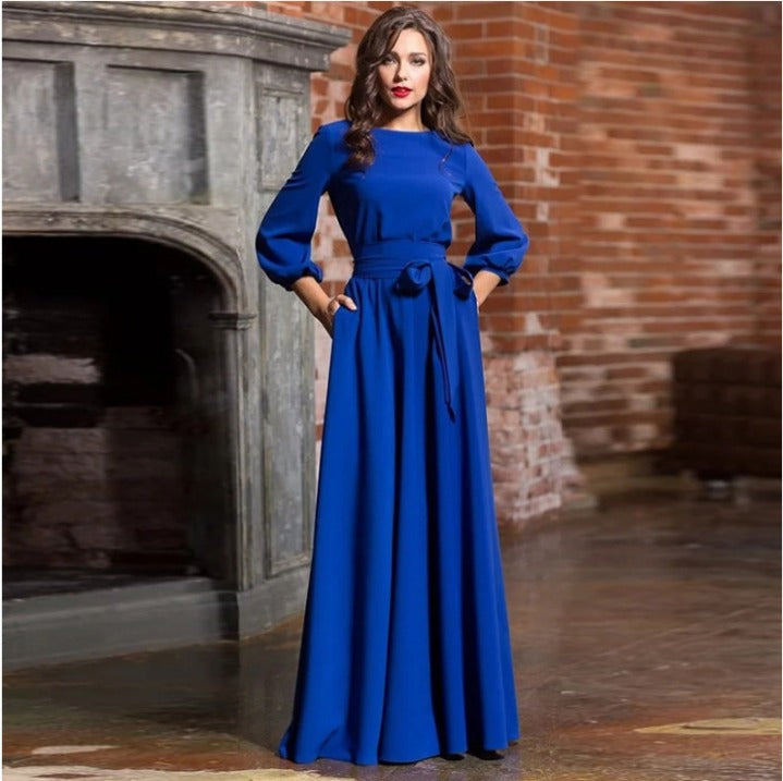 Navy Blue - A woman posing in a floor-length white crepe maxi dress featuring a boat neckline, three-quarter balloon sleeves, and a self-tie fabric sash at the waist, set against a modern interior backdrop.