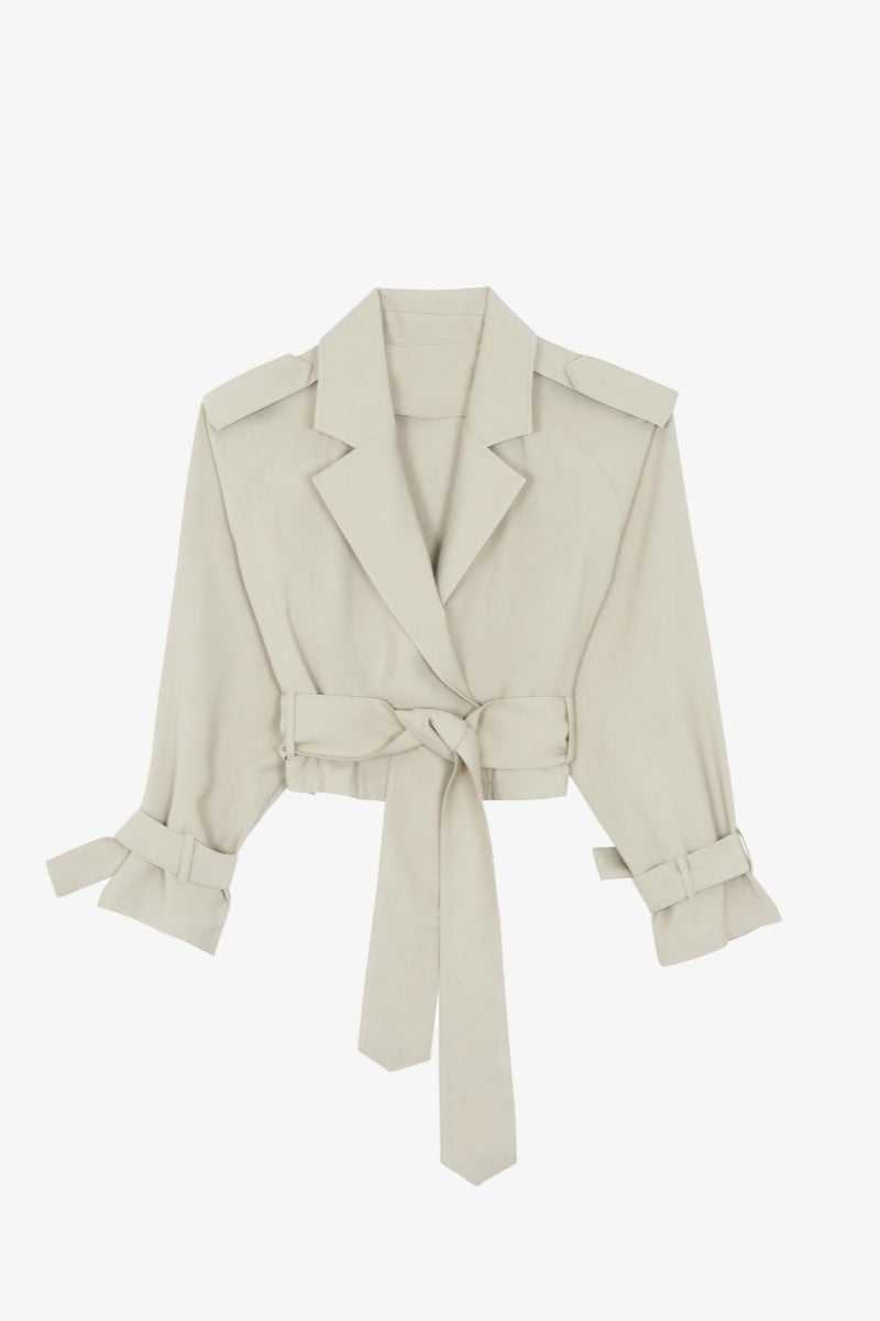 Cement - A model wearing a light beige cropped trench jacket with a belted waist, wide lapels, shoulder epaulettes, and relaxed sleeves.