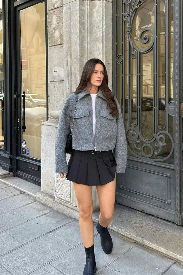 A woman wearing a women's grey textured cropped jacket with a silver front zipper and chest flap pockets, styled over a white t-shirt and black pleated mini skirt.