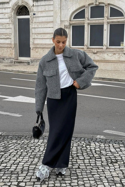 A woman wearing a women's grey textured cropped jacket with a silver front zipper and chest flap pockets, styled over a white t-shirt and black pleated mini skirt.