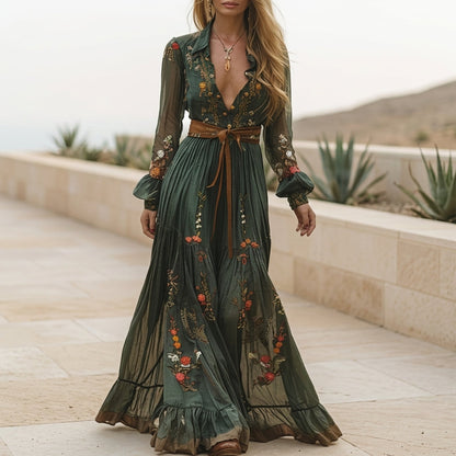 A woman wearing a dark green embroidered chiffon maxi dress with long balloon sleeves, a collared deep V-neckline, and a tiered pleated skirt, accessorised with a brown tie belt.