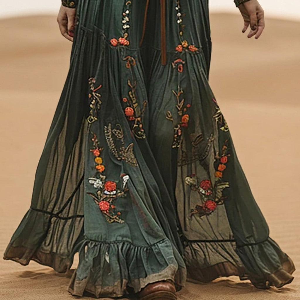 A woman wearing a dark green embroidered chiffon maxi dress with long balloon sleeves, a collared deep V-neckline, and a tiered pleated skirt, accessorised with a brown tie belt.